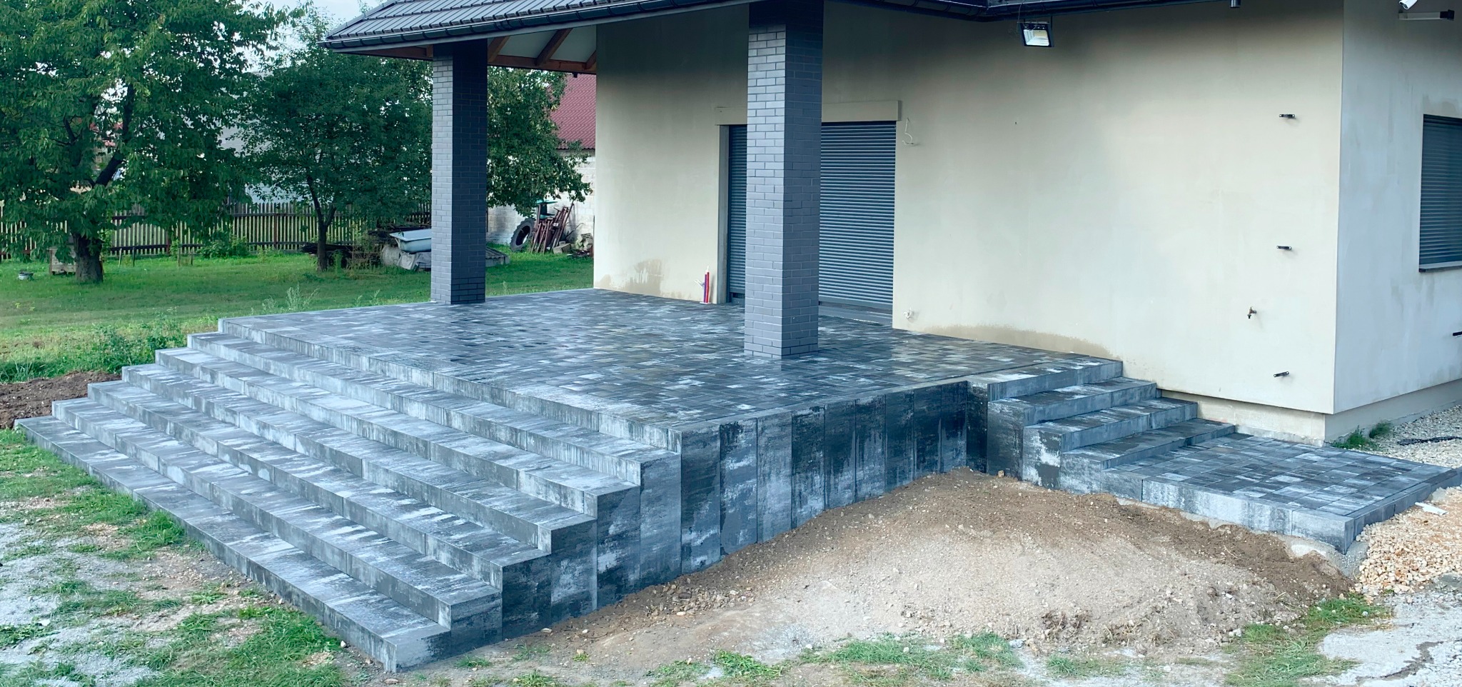 Nowo ułożony taras z kostki brukowej z schodami prowadzącymi do domu, widoczny fragment ogrodu i elewacja budynku.