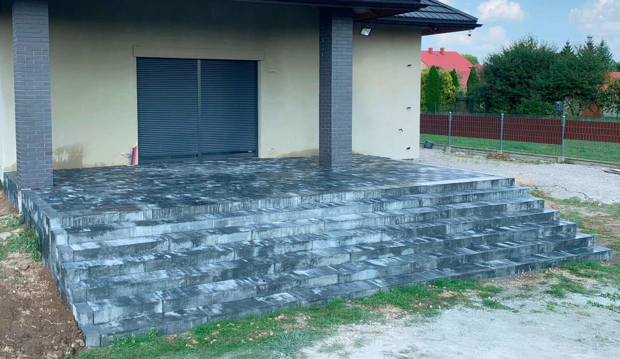 Schody i taras wyłożone szarą kostką brukową, z widoczną strukturą i zaciekami, prowadzące do budynku z zamkniętymi roletami i ceglanymi filarami, w tle ogrodzenie i zieleń.