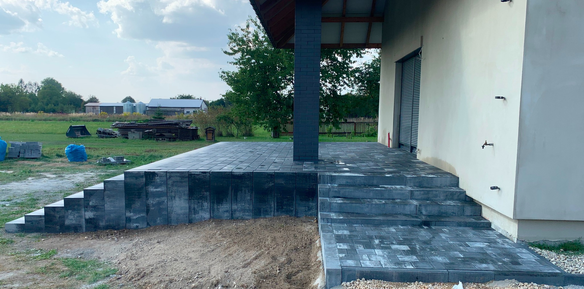 Nowo ułożony taras z ciemnoszarej kostki brukowej z niskimi schodami i filarem, widok na wiejski krajobraz w tle.