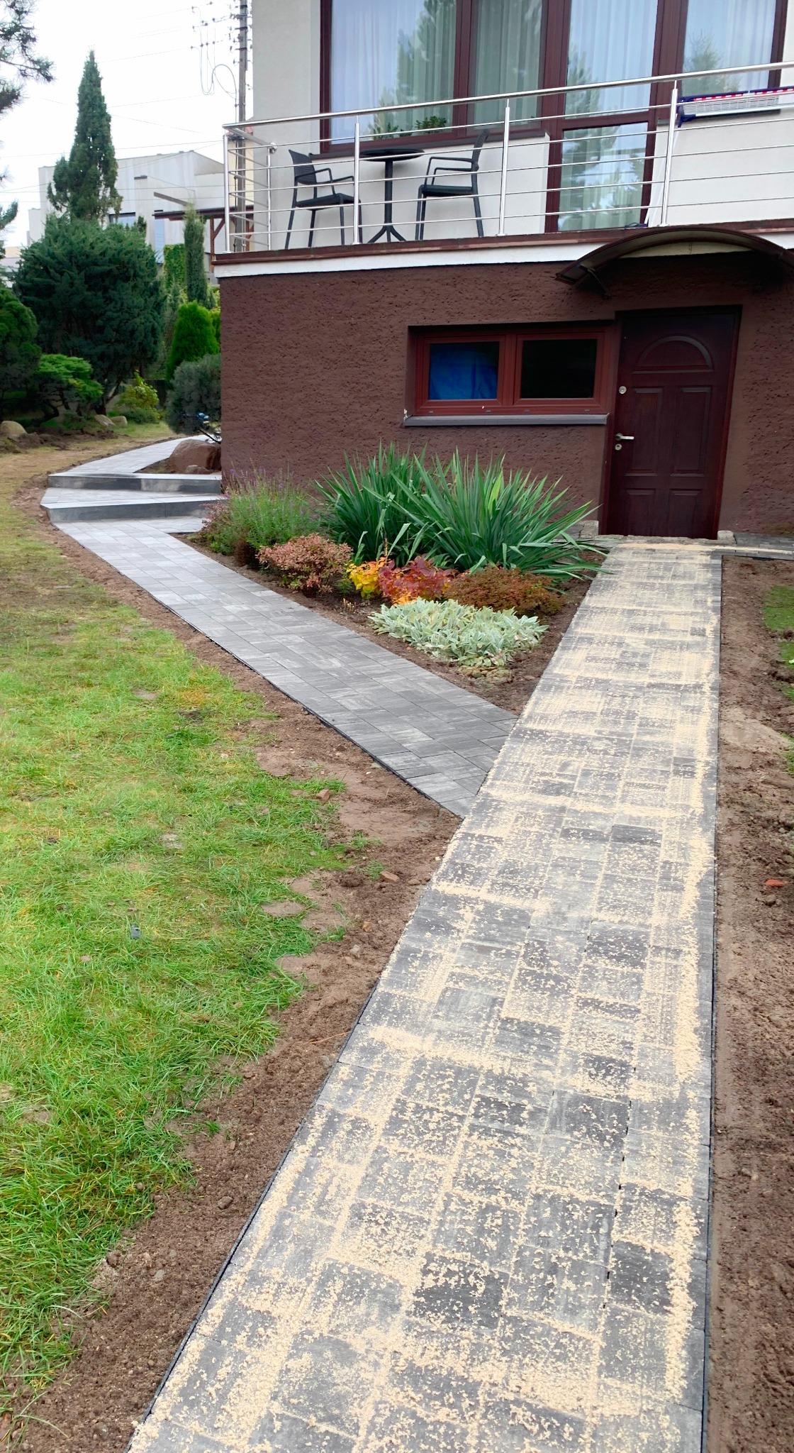 Świeżo ułożony chodnik z kostki brukowej w odcieniach szarości, posypany piaskiem, prowadzący do wejścia domu z brązową elewacją i balkonem z metalową balustradą.