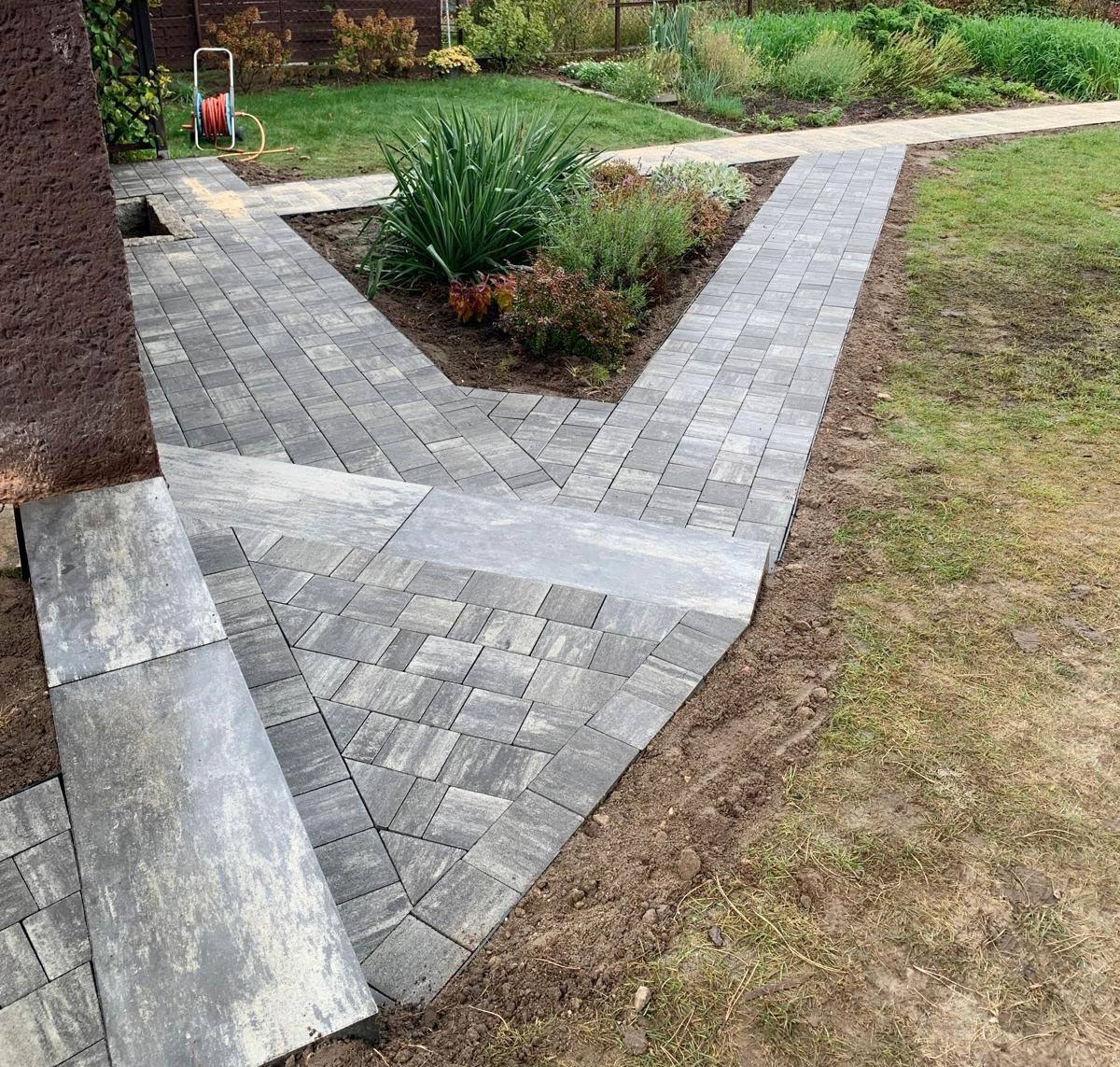 Ułożona ścieżka z szarej kostki brukowej prowadząca przez ogród, widoczne schody z kostki i rabata z roślinami ozdobnymi.
