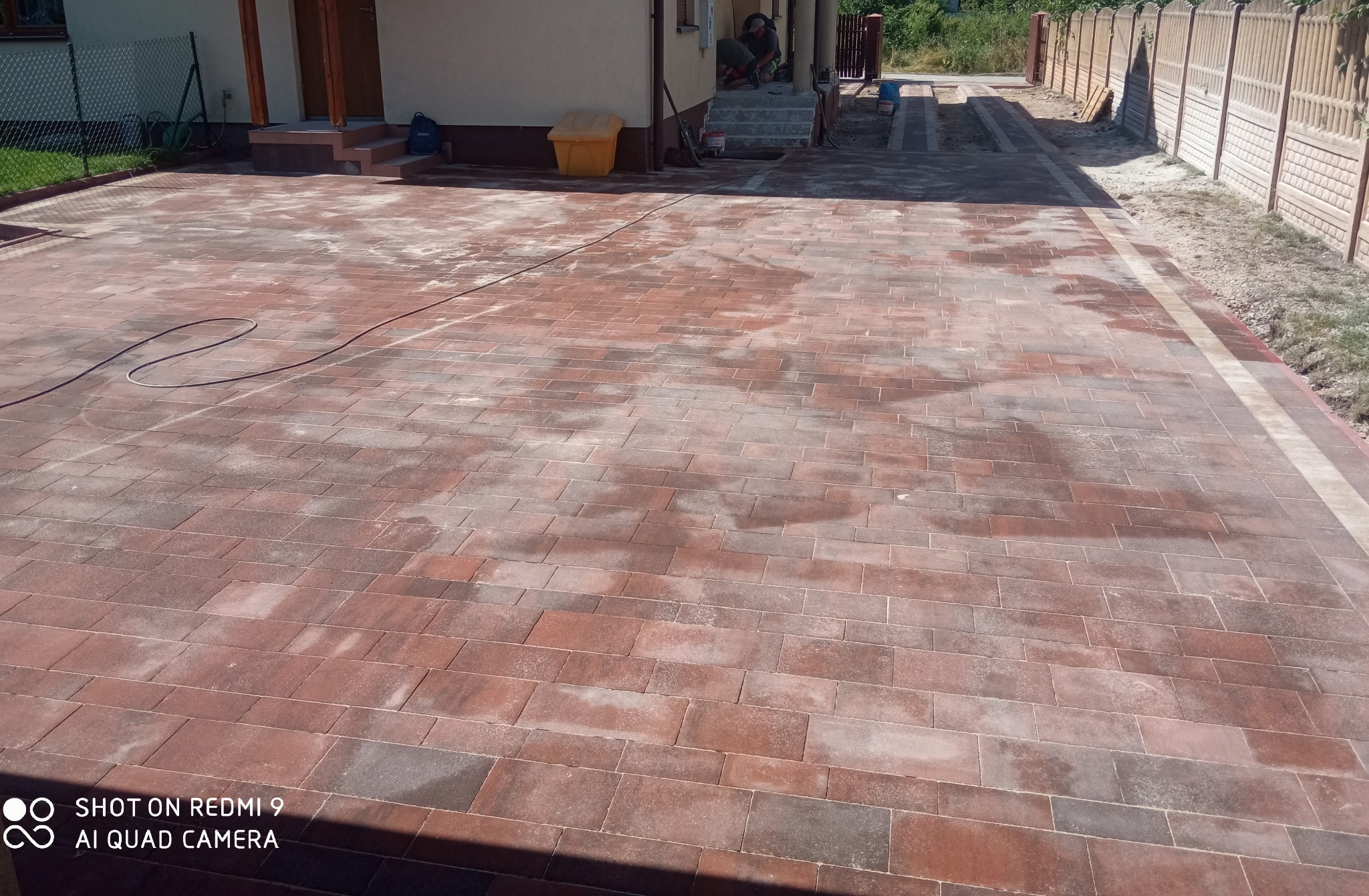 Nowo ułożona, czerwono-brązowa kostka brukowa na podjeździe, z widocznym obrzeżem i fragmentem domu w tle, częściowo mokra po myciu, z pozostawionym wężem ogrodowym.