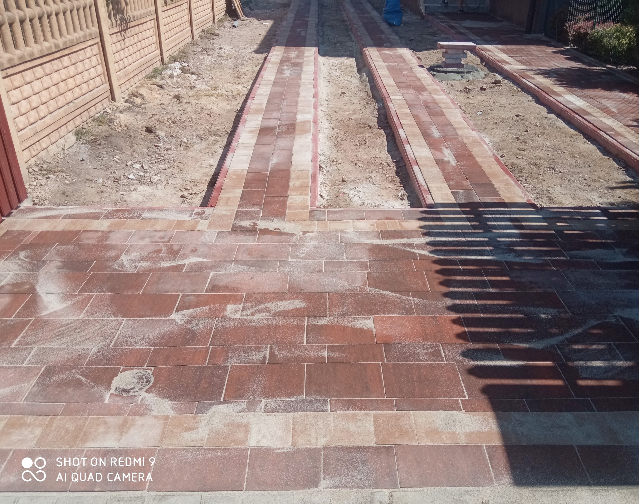 Nowo ułożona, czerwono-beżowa kostka brukowa na podjeździe, z widocznymi śladami piasku do wypełniania szczelin, oraz przygotowane koryta na trawniki po bokach.