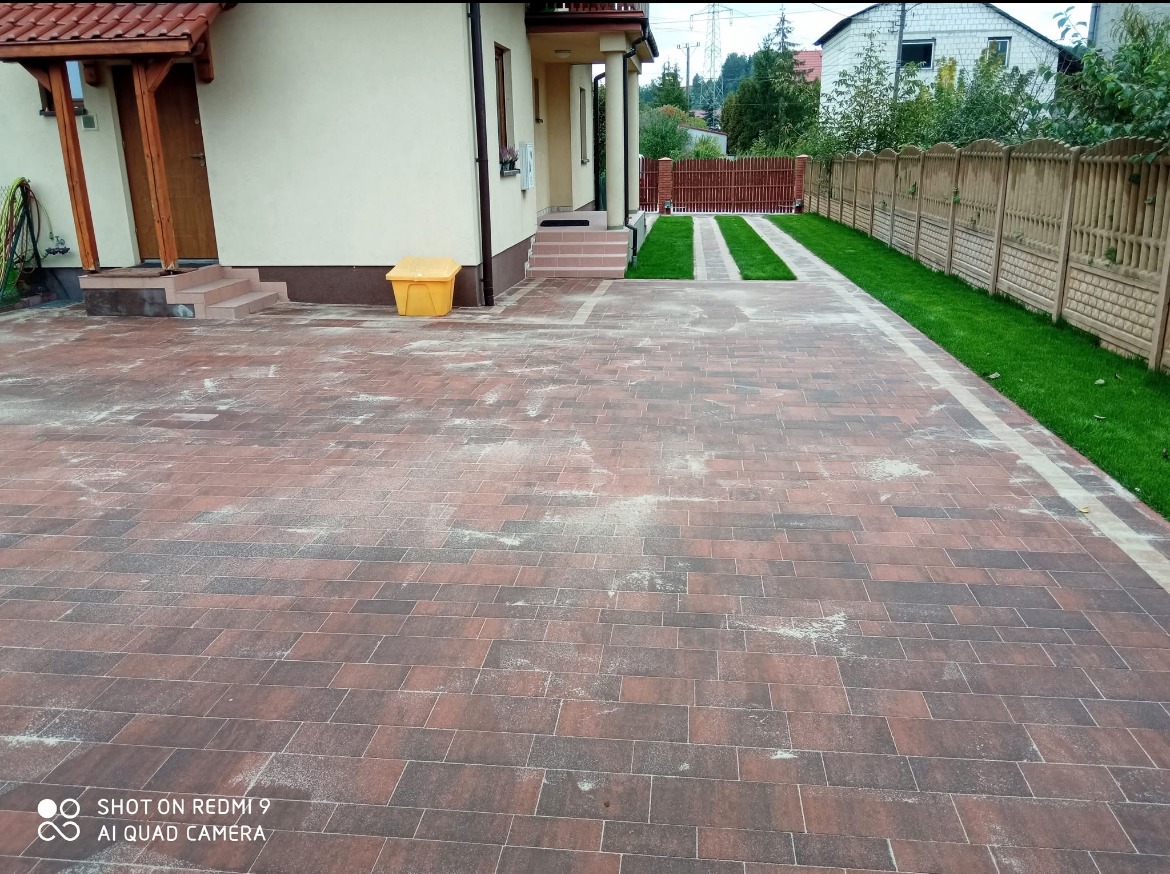 Nowo ułożona, brązowo-szara kostka brukowa na podjeździe, z dwoma pasami trawy pośrodku i obrzeżem z trawy po bokach, żółty pojemnik na piasek przy budynku.