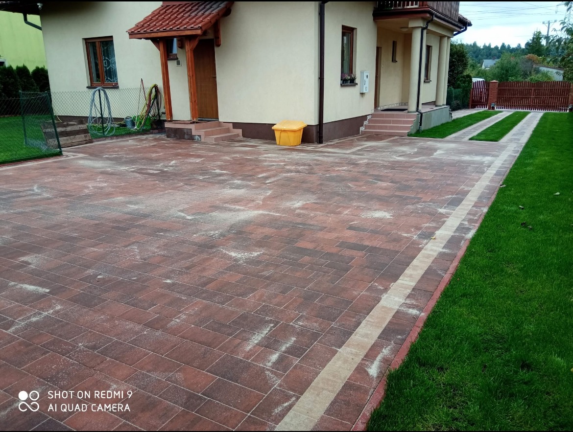 Rozległy podjazd z czerwono-brązowej kostki brukowej ułożonej przed domem z beżową elewacją, widoczne ślady piasku na powierzchni, obrzeża z jasnej kostki, trawnik po prawej stronie i pasy trawy...