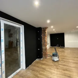 Wnętrze w trakcie remontu: położone panele podłogowe, pomalowana na czarno ściana, fragment ściany z cegły, odkurzacz przemysłowy i okno z folią ochronną.