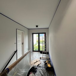 Jakub Ponc - Korytarz w trakcie odświeżania: świeżo pomalowane ściany w odcieniach szarości zabezpieczone niebieską taśmą malarską przy suficie, podłoga przykryta folią ochronną, widoczna drabina aluminiowa...