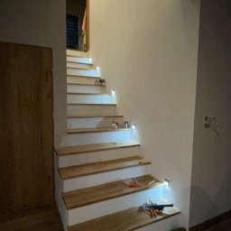 Jakub Ponc - Drewniane schody z białymi podstopnicami i wbudowanym oświetleniem LED, widoczne narzędzia i kable na dolnym stopniu, drzwi w tle.