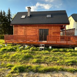 Jakub Ponc - Drewniany domek z ciemną dachówką i tarasem, podniesiony na betonowych blokach, otoczony trawiastym terenem z kałużami, widziany w słoneczny dzień.