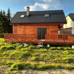 Drewniany domek z ciemną dachówką i tarasem, podniesiony na betonowych blokach, otoczony trawiastym terenem z kałużami, widziany w słoneczny dzień.