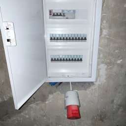 Jakub Ponc - Otwarta, biała skrzynka elektryczna z bezpiecznikami na szarej, surowej ścianie, poniżej widoczny kabel z czerwono-białą końcówką i worek z odpadami budowlanymi.