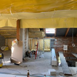 Jakub Ponc - Poddasze w trakcie prac w Brennej: widoczne ocieplenie folią, wełna mineralna, fragmenty murowanej ściany, instalacja elektryczna, drabina i narzędzia pracy.