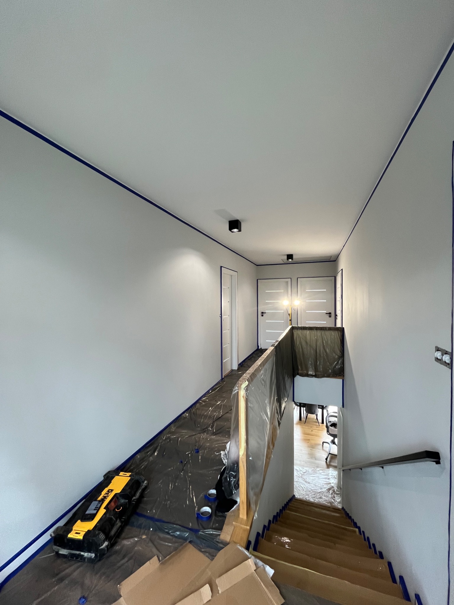 Świeżo pomalowany korytarz z zabezpieczonymi powierzchniami taśmą malarską i folią, widoczny laserowy niwelator DeWalt, przygotowany do dalszych prac wykończeniowych.