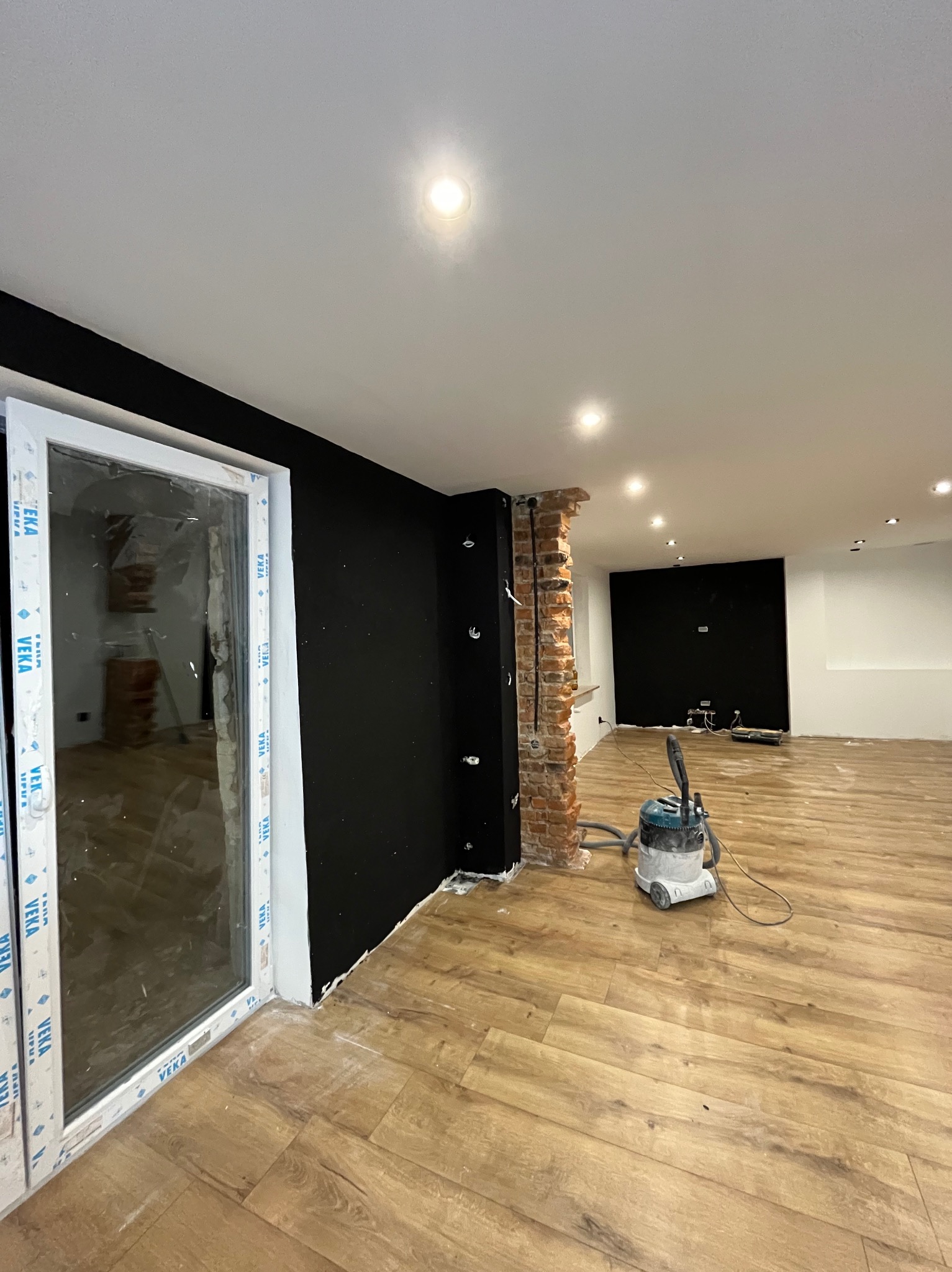 Wnętrze w trakcie remontu: położone panele podłogowe, pomalowana na czarno ściana, fragment ściany z cegły, odkurzacz przemysłowy i okno z folią ochronną.