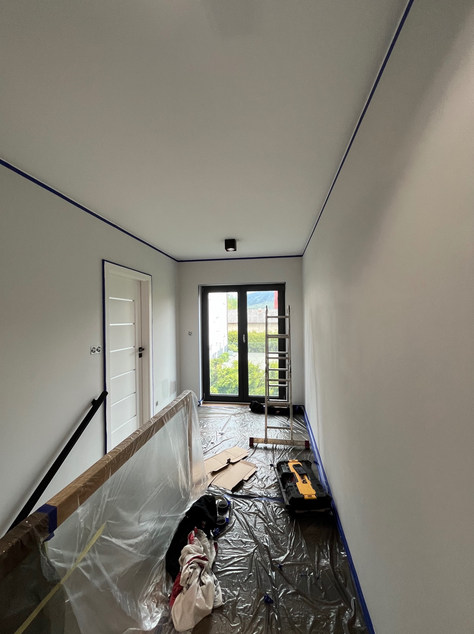Korytarz w trakcie odświeżania: świeżo pomalowane ściany w odcieniach szarości zabezpieczone niebieską taśmą malarską przy suficie, podłoga przykryta folią ochronną, widoczna drabina aluminiowa...