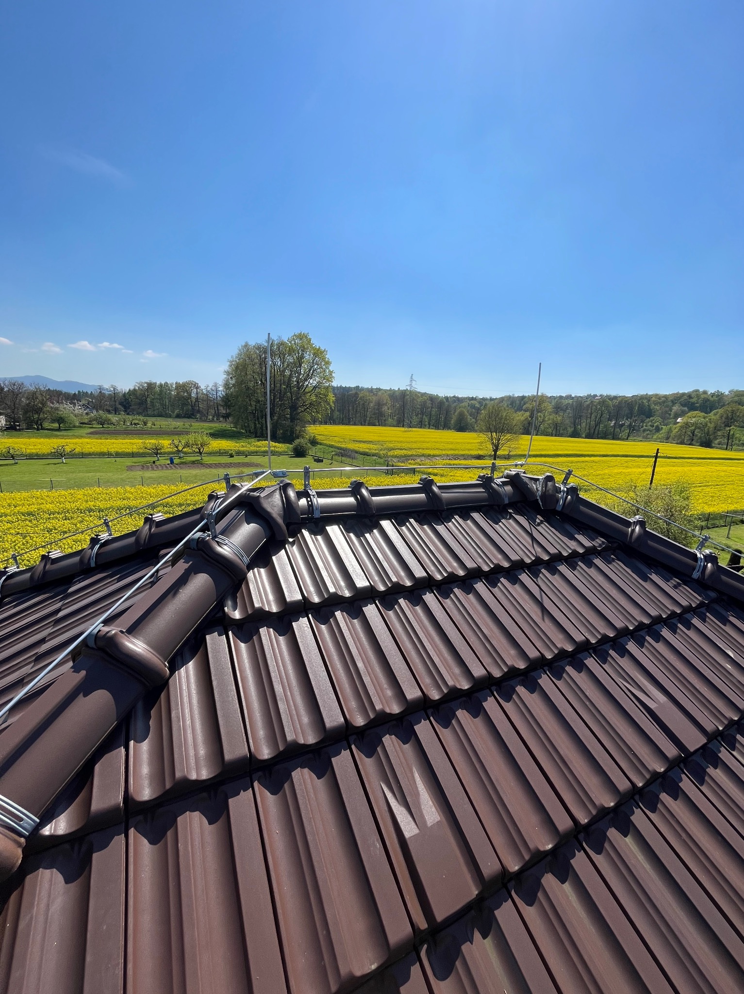 Widok z góry na brązowy dach z dachówki ceramicznej z zamontowanym systemem odgromowym na tle kwitnącego na żółto pola rzepaku i błękitnego nieba.