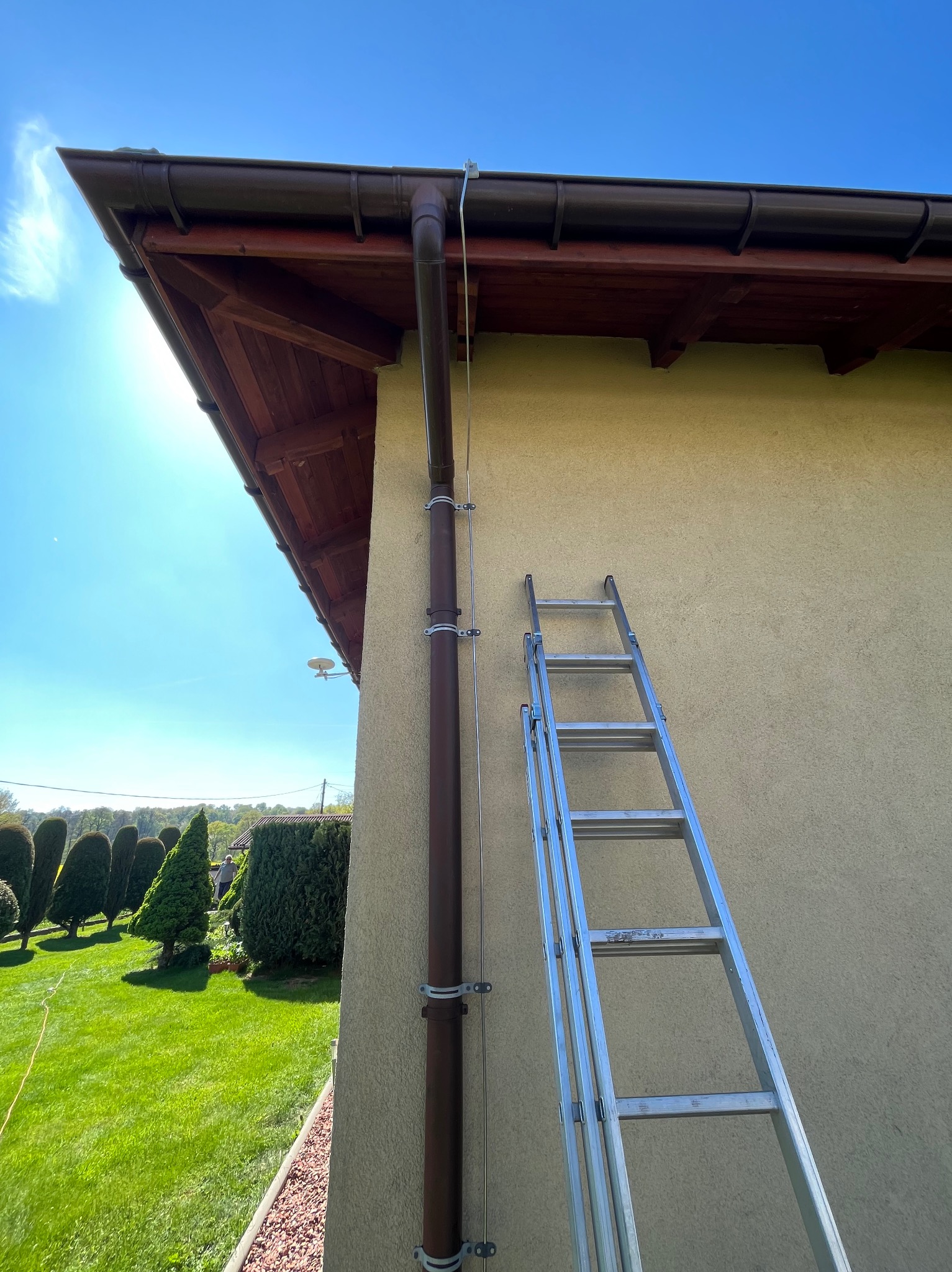 Brązowa rynna spustowa zamontowana na elewacji budynku, zabezpieczona metalowymi obejmami, obok oparta aluminiowa drabina, widok na zadbany ogród z zielonym trawnikiem i ozdobnymi tujami.