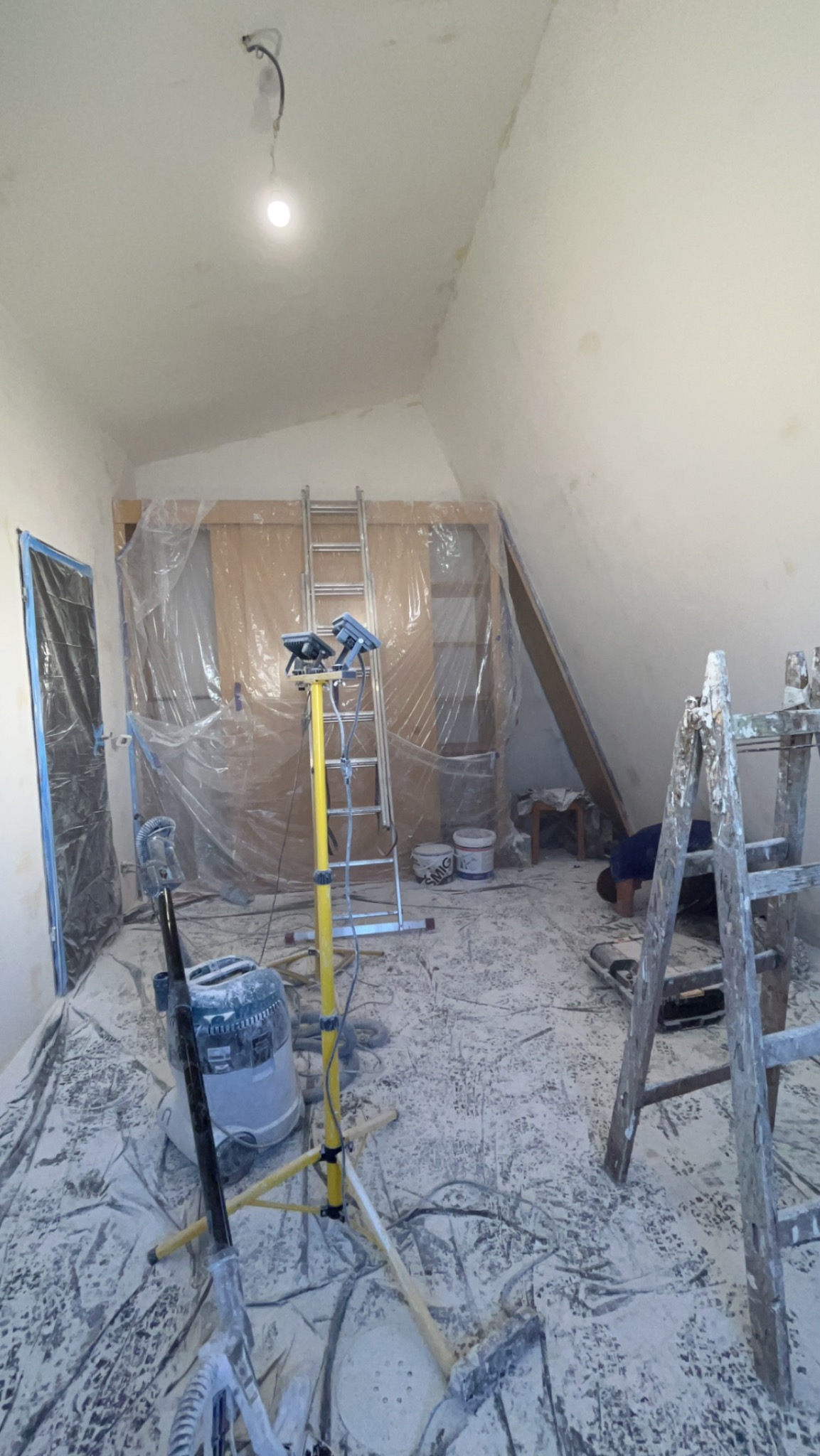 Poddasze w trakcie prac wykończeniowych: pył gipsowy na podłodze zabezpieczonej folią, drabiny, lampa budowlana na statywie, odkurzacz przemysłowy, zakryte folią drzwi i fragment ściany skośnej.