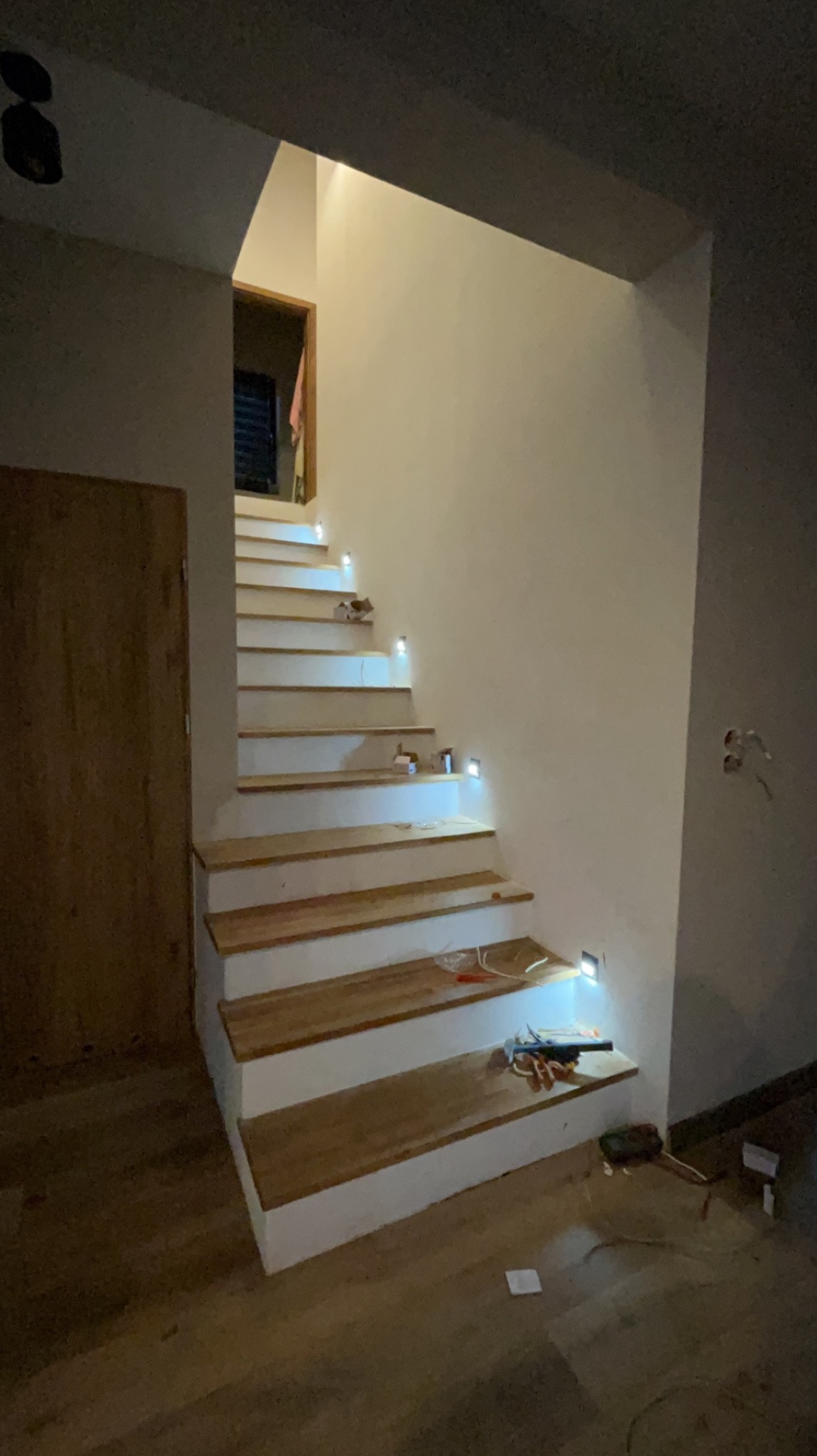 Drewniane schody z białymi podstopnicami i wbudowanym oświetleniem LED, widoczne narzędzia i kable na dolnym stopniu, drzwi w tle.