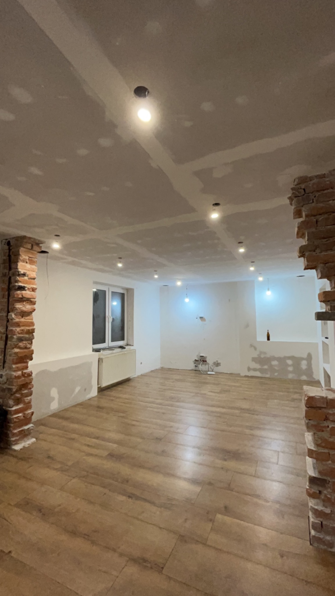 Pokój po remoncie: surowy strop z płyt kartonowo-gipsowych ze śladami szpachlowania, nowa podłoga z paneli, fragment ściany z cegły, instalacja elektryczna w trakcie montażu.