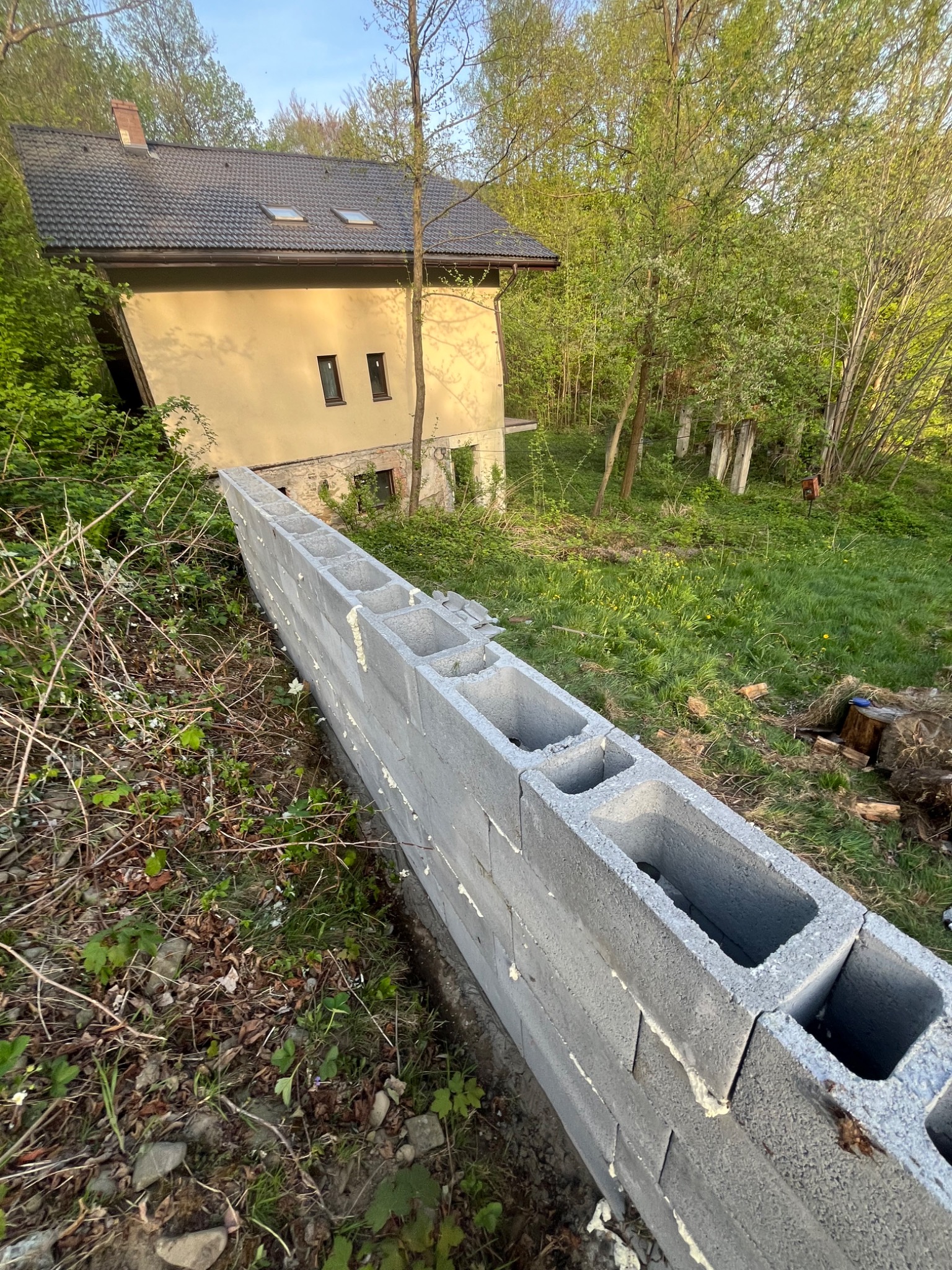 Wznoszenie ściany z szarych bloczków betonowych na nierównym terenie z widokiem na dom w tle, widać świeży biały klej między blokami.