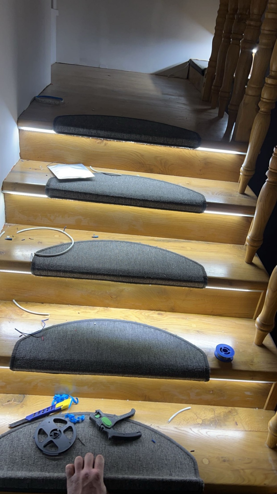 Montaż dywanowych nakładek na drewnianych schodach z podświetleniem LED, widoczne narzędzia do cięcia kabli i fragment stopy