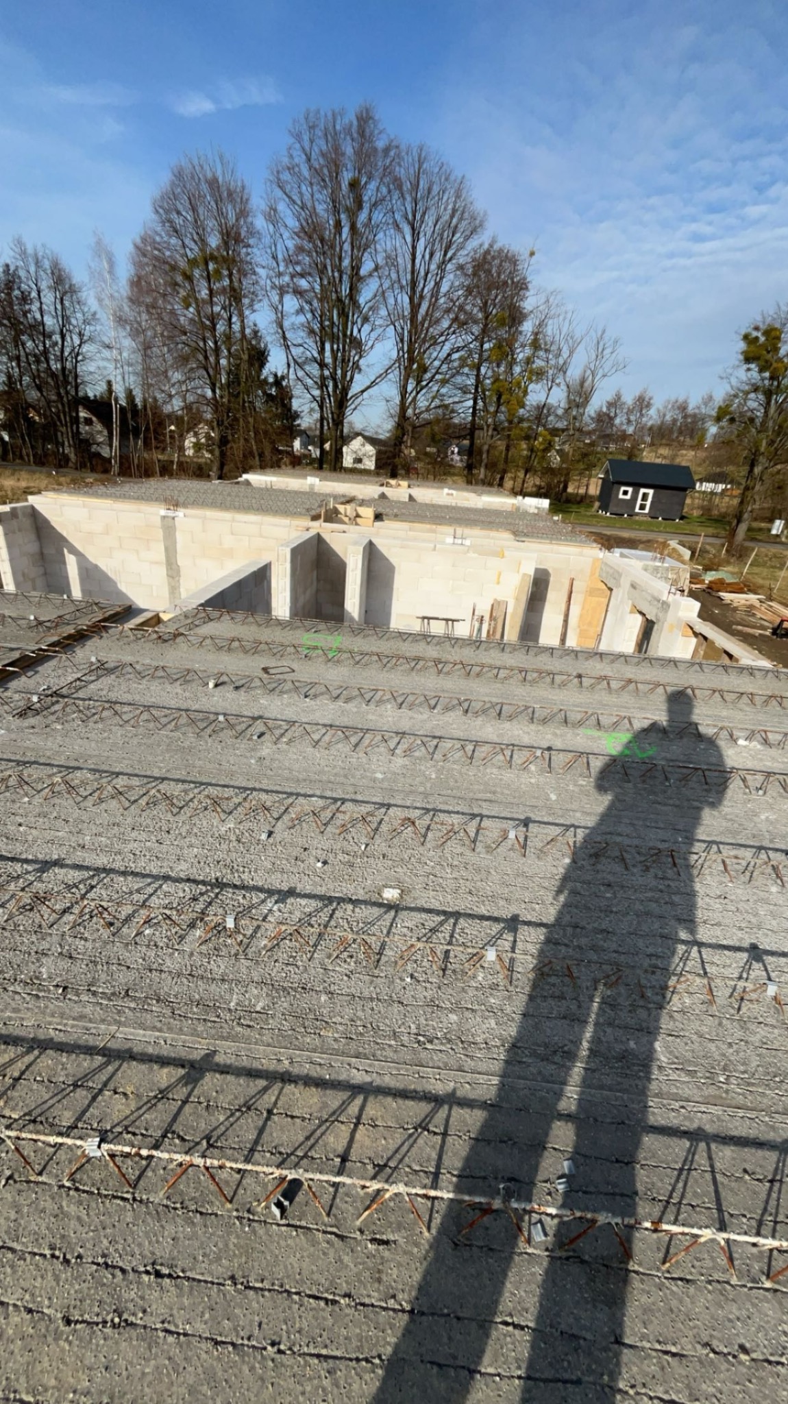 Fundamenty domu jednorodzinnego w trakcie budowy, widoczne zbrojenia i ściany fundamentowe z bloczków betonowych, cień osoby fotografującej na pierwszym planie.