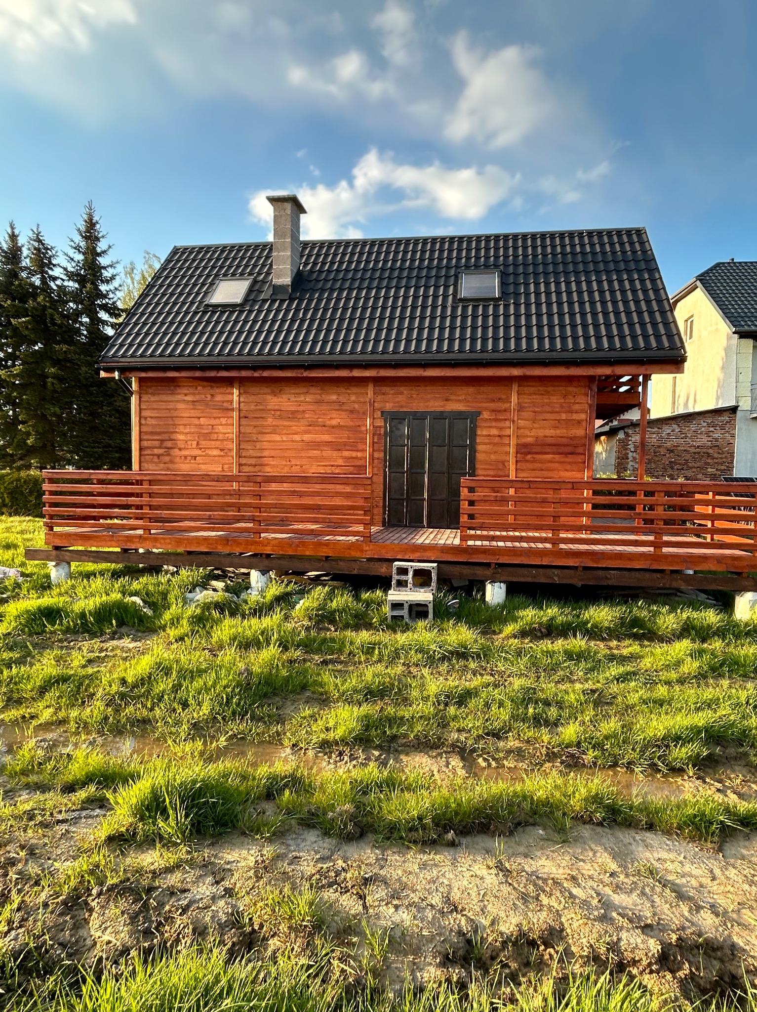 Drewniany domek z ciemną dachówką i tarasem, podniesiony na betonowych blokach, otoczony trawiastym terenem z kałużami, widziany w słoneczny dzień.