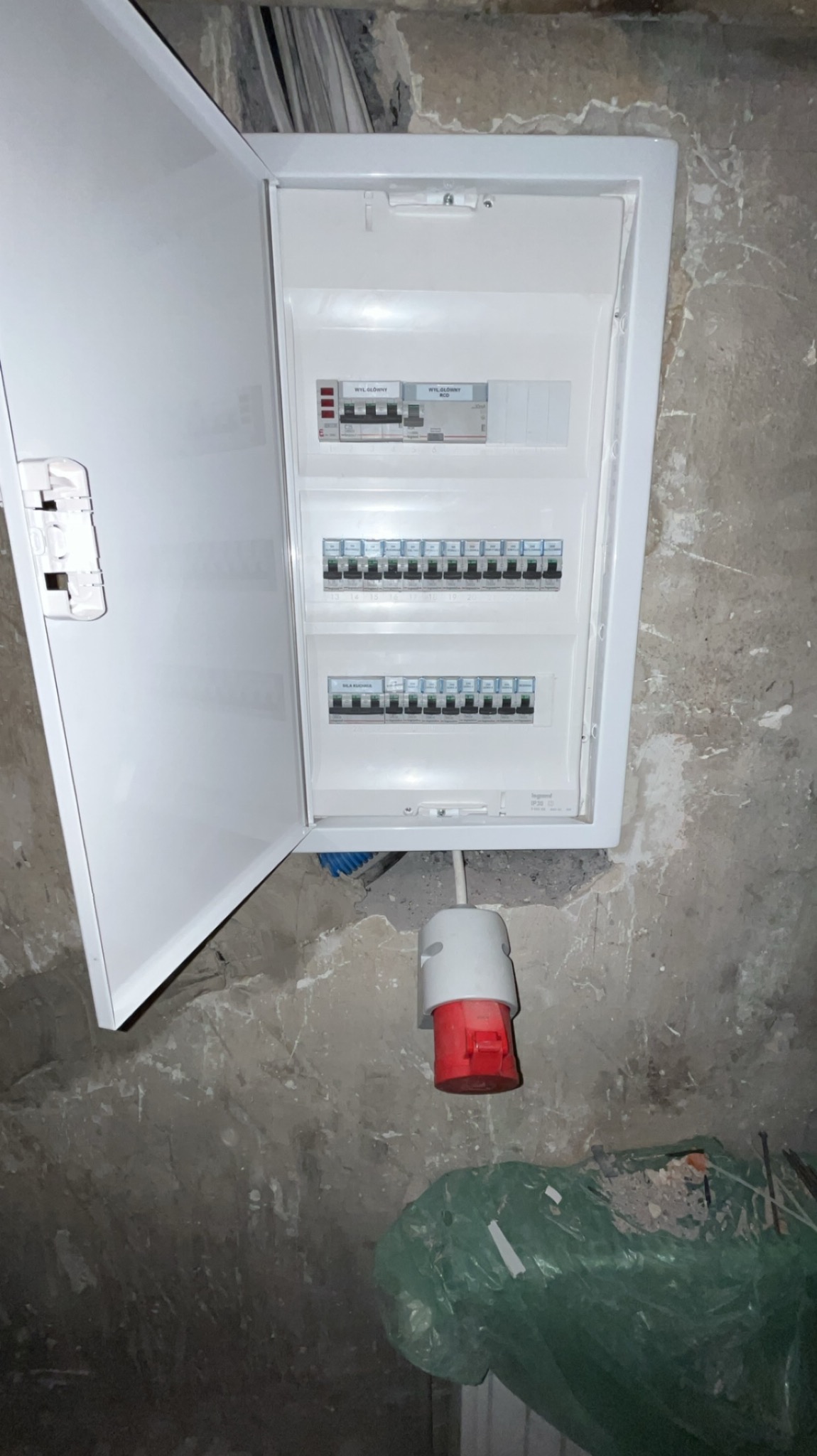 Otwarta, biała skrzynka elektryczna z bezpiecznikami na szarej, surowej ścianie, poniżej widoczny kabel z czerwono-białą końcówką i worek z odpadami budowlanymi.