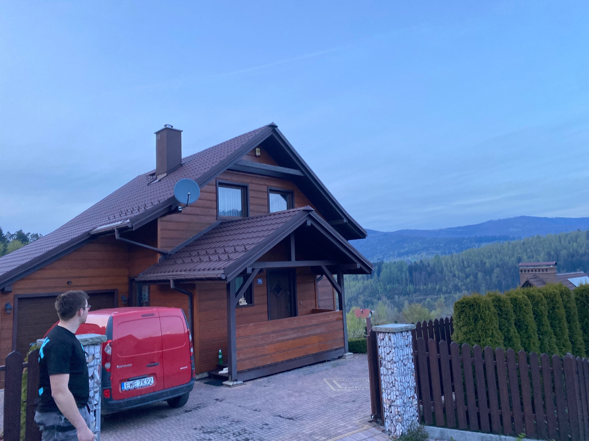 Drewniany dom jednorodzinny z czerwoną furgonetką zaparkowaną przed garażem, widok na góry w tle, pochmurne niebo.