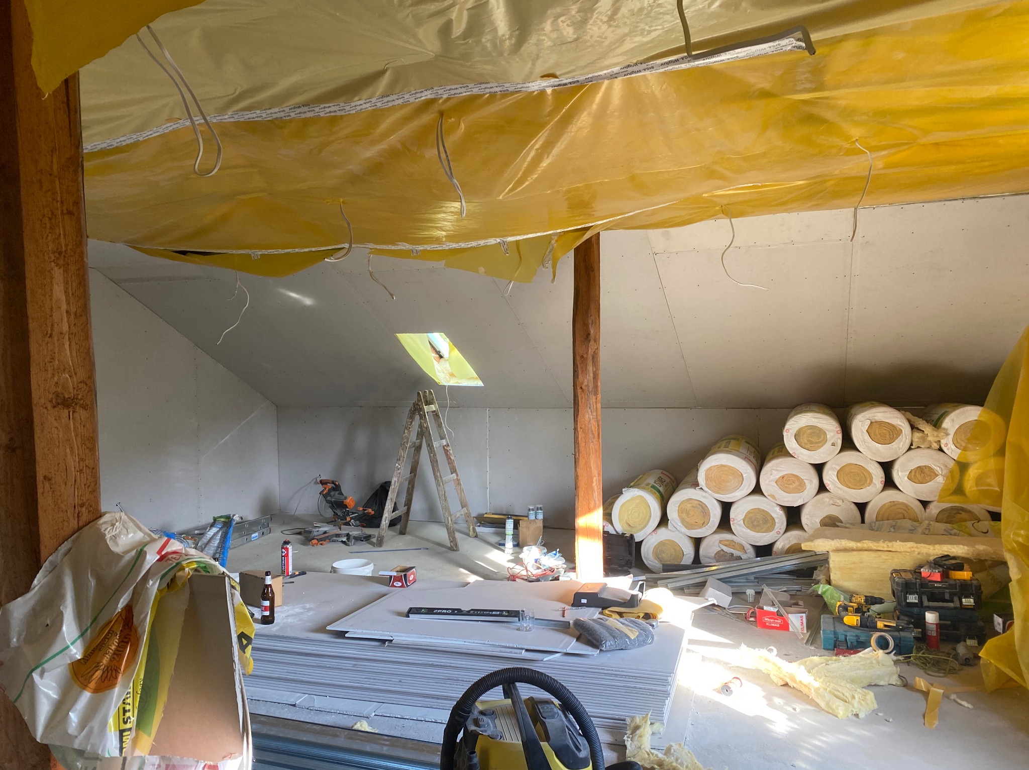 Poddasze w trakcie ocieplania i montażu płyt karton gips, widoczne rolki wełny mineralnej, płyty gipsowe, drabina i narzędzia, folia paroizolacyjna pod dachem.