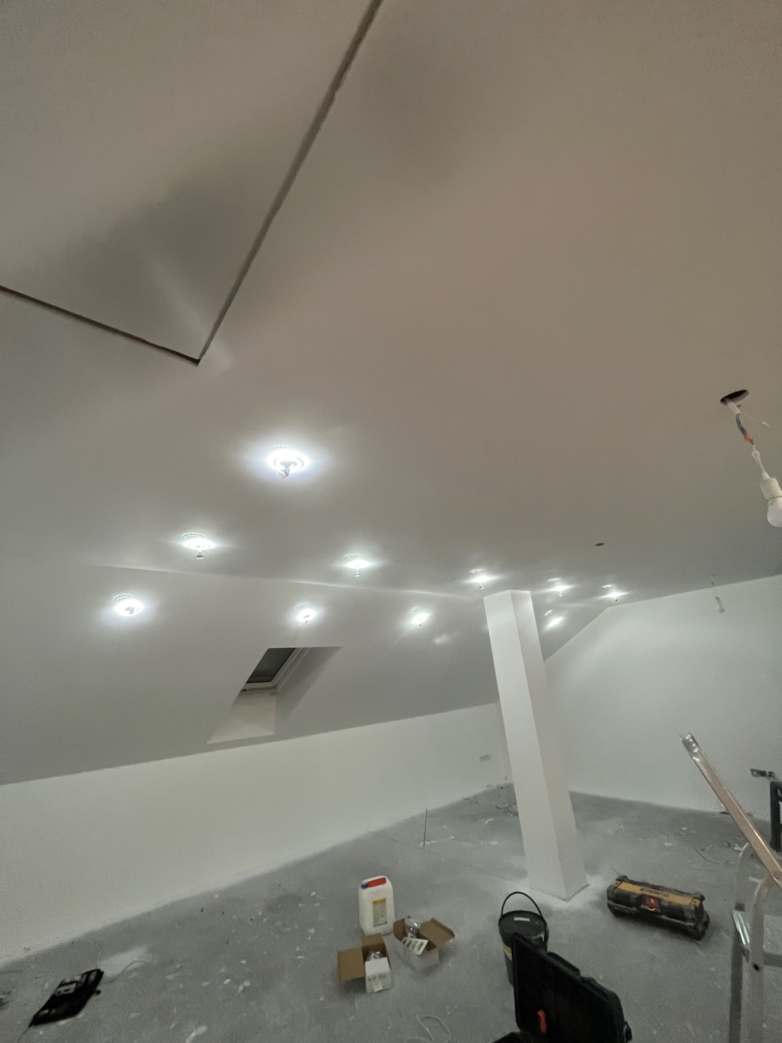 Poddasze po remoncie: białe ściany, sufit ze świetlikami i zamontowanymi punktowymi lampami, widoczne narzędzia i materiały budowlane na podłodze.