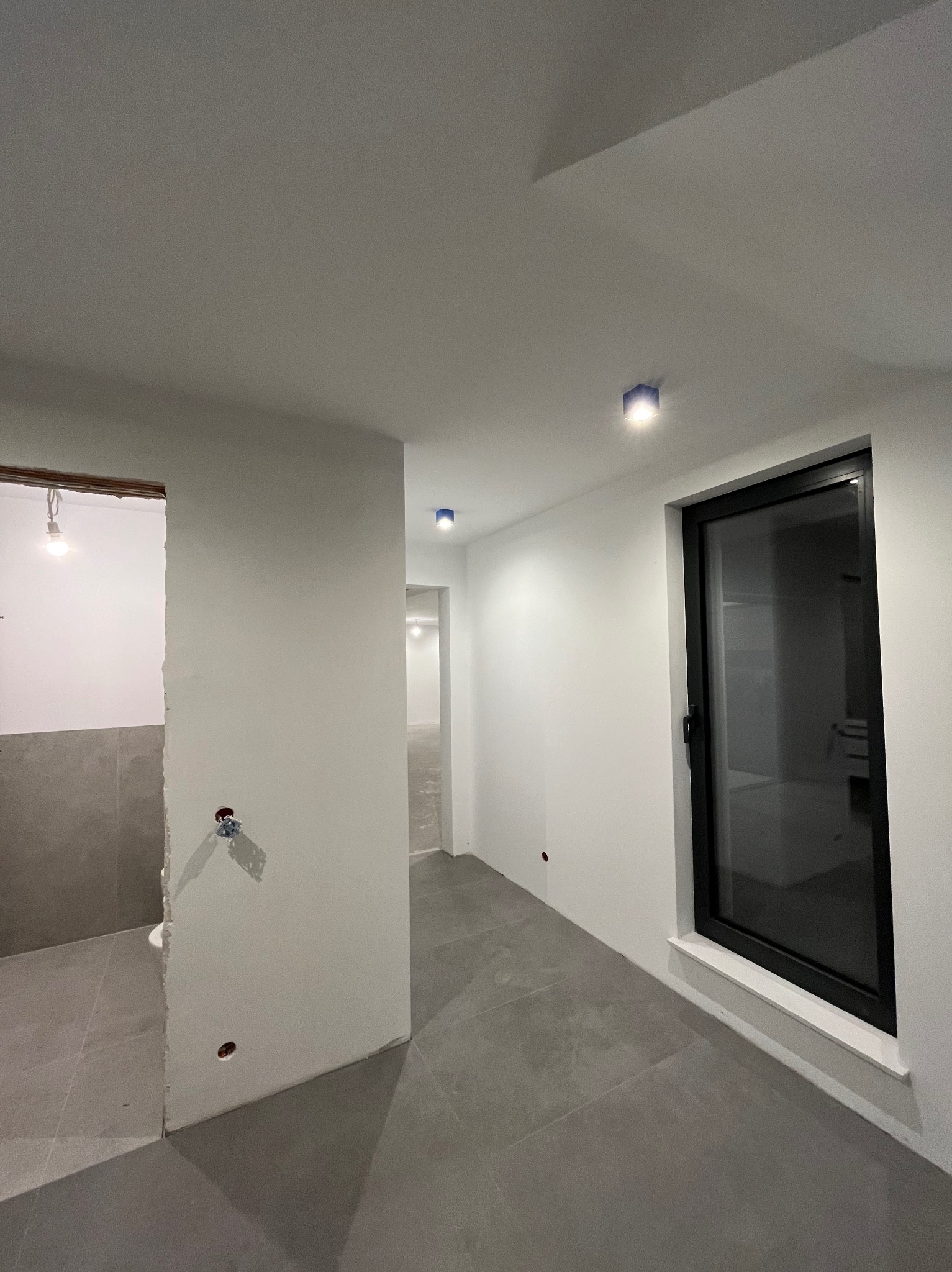 Wnętrze w trakcie remontu: szare płytki podłogowe, białe ściany z widocznymi punktami elektrycznymi, czarna rama okienna i sufit z oświetleniem punktowym.