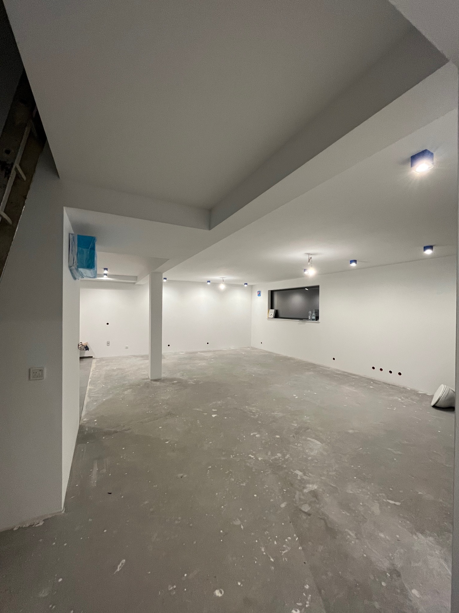 Przestronne, jasne wnętrze w trakcie remontu: surowy beton na podłodze, świeżo pomalowane białe ściany i sufit z punktowym oświetleniem LED. Widoczne zabezpieczenia folią malarską. Stan surowy...