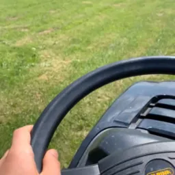 Widok z perspektywy operatora na maszynę do koszenia trawy marki Alpina, w trakcie koszenia rozległego trawnika z drzewami w tle.