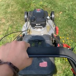 Szymon Usługi ogrodnicze - Osoba z bransoletką na nadgarstku trzyma czarny uchwyt kosiarki spalinowej nad trawnikiem z żółtymi kwiatami.