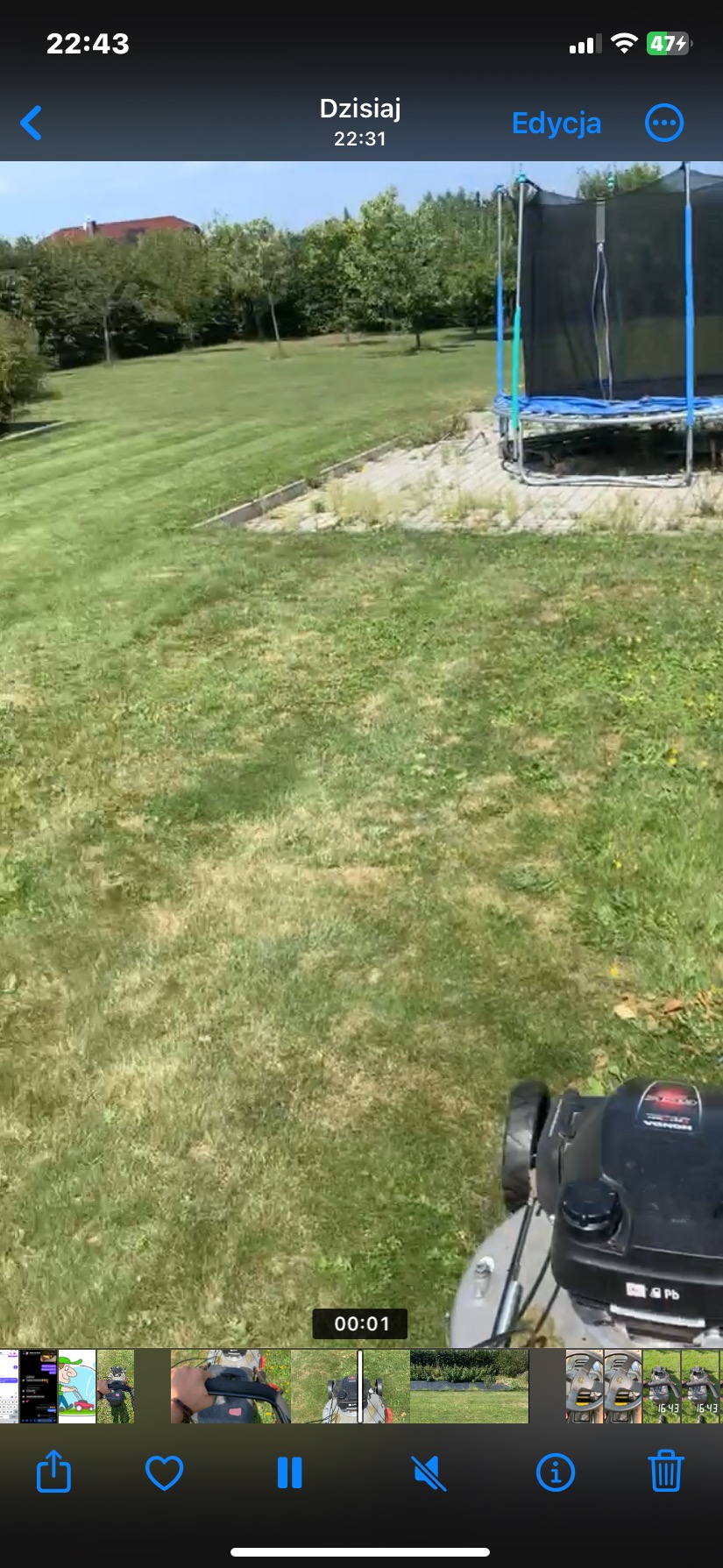 Kosiarka spalinowa w trakcie koszenia trawy na nierównym terenie, widoczny trawnik przed domem i trampolina w tle.