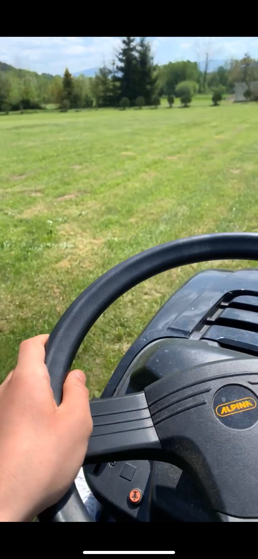 Widok z perspektywy operatora na maszynę do koszenia trawy marki Alpina, w trakcie koszenia rozległego trawnika z drzewami w tle.