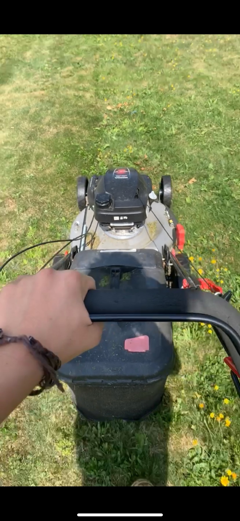 Osoba z bransoletką na nadgarstku trzyma czarny uchwyt kosiarki spalinowej nad trawnikiem z żółtymi kwiatami.