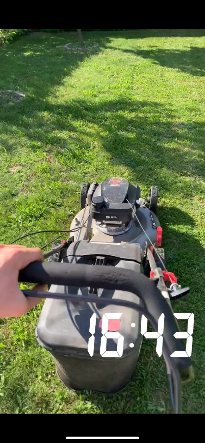 Osoba z perspektywy pierwszej osoby prowadzi kosiarkę spalinową po trawniku w słoneczny dzień. Widoczny kosz na trawę i fragment trawnika.