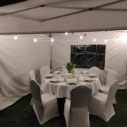 Ustawiony okrągły stół z białym obrusem i nakryciami dla sześciu osób, otoczony krzesłami w białych pokrowcach, pod białym namiotem oświetlonym girlandą żarówek, z widoczną czarną folią w oknie...