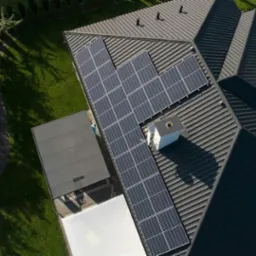 Widok z drona na ciemny dach z panelami słonecznymi w Przasnyszu, sąsiadujący z zielonym trawnikiem i drzewami.