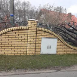 Żółty mur z cegły klinkierowej z dekoracyjnym, metalowym ogrodzeniem imitującym gałęzie drzew, wbudowane białe drzwi rewizyjne, widok z poziomu ulicy.