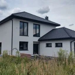 Nowoczesny, dwukondygnacyjny dom z garażem, wykończony na biało z czarną dachówką, otoczony wysoką trawą, w tle pochmurne niebo.