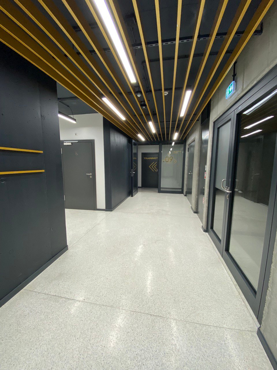 Wnętrze nowoczesnego biura: korytarz z ciemnymi ścianami, jasną podłogą z lastryko i sufitem z drewnianymi belkami i oświetleniem liniowym LED.