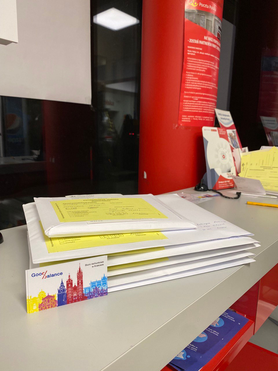 Stos białych kopert z żółtymi naklejkami adresowymi leżący na biurku, obok wizytówka biura rachunkowego z kolorową panoramą Krakowa.