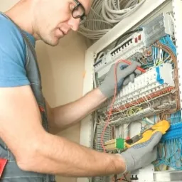 Elektryk w okularach, w szarych rękawicach, mierzy napięcie w rozdzielnicy elektrycznej za pomocą multimetru; widoczne kable i przewody.