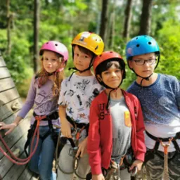 Grupa czworga dzieci w kaskach i uprzężach, stojąca przed ścianką wspinaczkową w parku linowym, gotowa do rozpoczęcia aktywności.