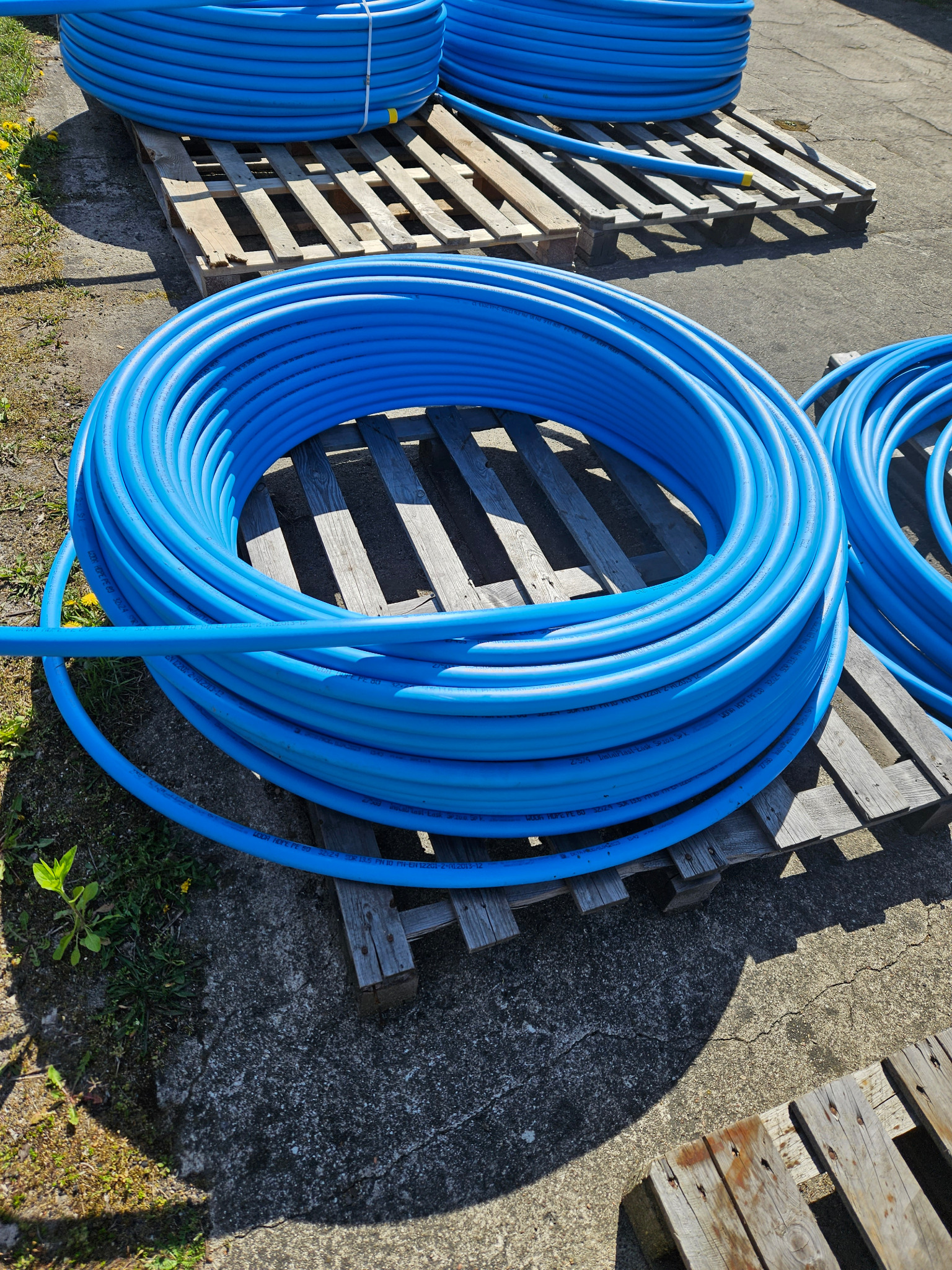 Zwoje niebieskich rur HDPE na drewnianych paletach, widoczne napisy na rurach, teren zewnętrzny.