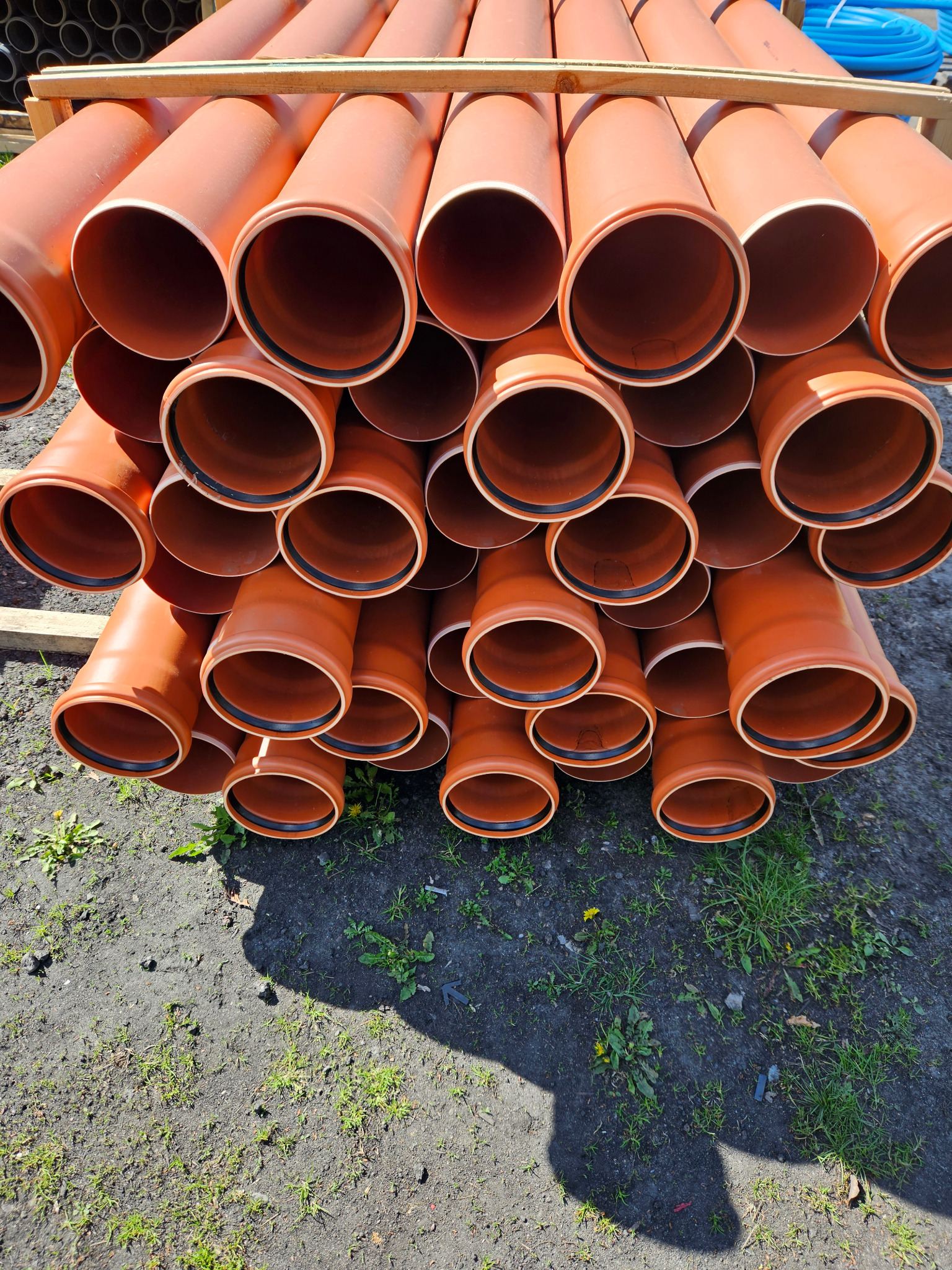 Stos pomarańczowych rur kanalizacyjnych z czarnymi uszczelkami, ułożonych na drewnianych belkach, rzucających cień na ziemię z trawą.